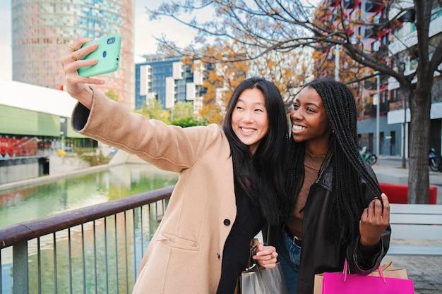 A couple of multiracial happy girls having fun takng a photo with a cellphone Two young women smiling doing a selfie portrait with a smart phone in a shopping sale day Lifestyle concept