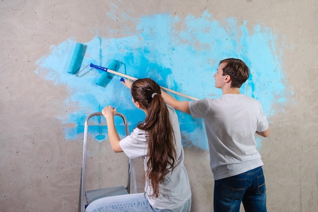 Couple in new home during repair works painting wall together happy family holding paint roller pain