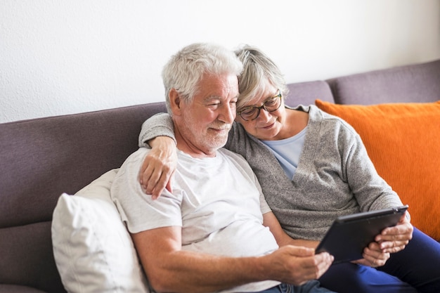 소파에 안긴 같은 태블릿을 보고 웃고 있는 노인 부부 - 실내, 가정 개념 - 백인들은 기술을 사용하여 성숙하고 은퇴한 남녀