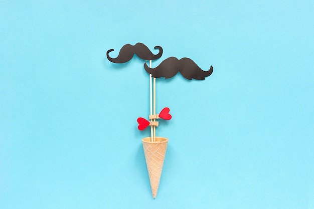 Couple paper mustache props on stick fastened clothespin heart in ice cream waffle cone on blue background