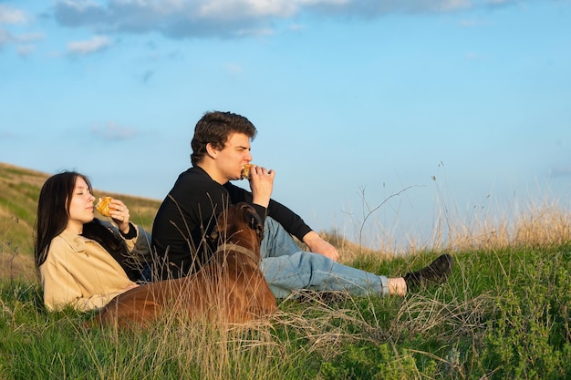 A couple of people in love with a big dog in nature sit on the ground and eat hamburgers