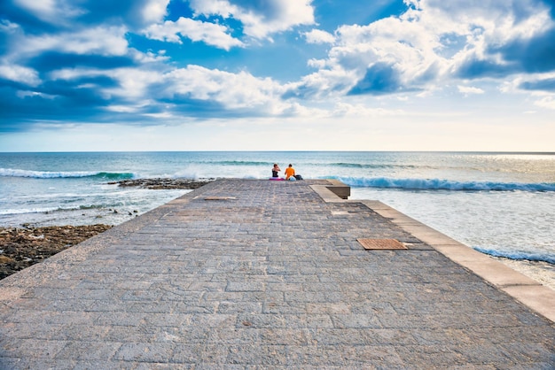 Couple people sitting on edge of stone pier looking at sea landscape