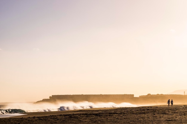 Couple of people traveler tourists walking together at the beach during a wave storm sunset golden light with big wave in background