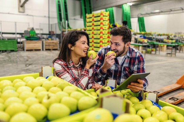 Couple in plaid shirts eat green apples and take selfie photos with a tablet in the warehouse