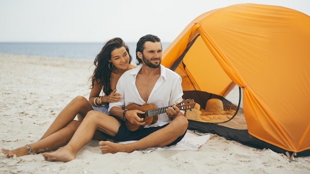 Coppia che suona l'ukulele con una tenda arancione sulla spiaggia