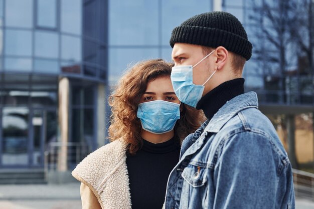 Couple in protective masks have a walk outdoors in the city near business building at quarantine time Conception of coronavirus