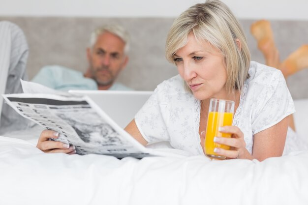 Couple reading newspaper and using laptop in bed