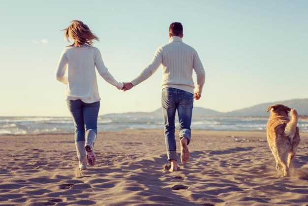 Photo couple running on the beach holding their hands with dog on autmun day