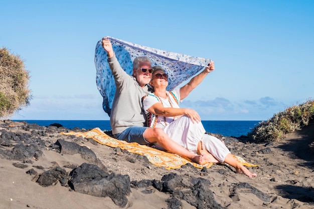 couple of senior caucasian hippy style and life rest on the rocky beach in tenerife. colors and alternative lifestyle for totally freedom concept near the ocean. alternative colorful clothes to live f