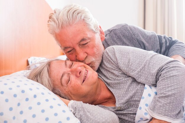 Couple of senior man and woman waking up and smiling with a hug while are in the bed at home. he kiss her with love for a life together