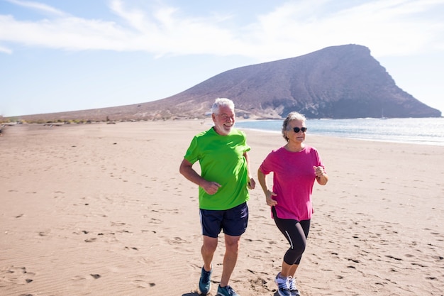 Coppia di anziani e persone mature che corrono e fanno jogging insieme sulla spiaggia sulla sabbia - stile di vita e concetto sano e fitness - pensionati attivi che si divertono e fanno esercizio