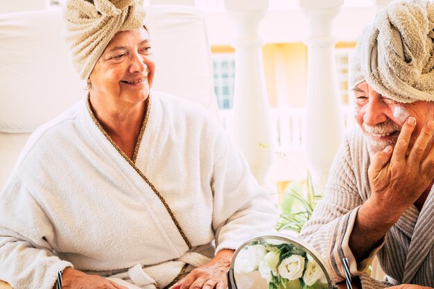 Couple of seniors sitting together at hotel with spa and beauty tratament - close up of man putting some cream on his face with her wife looking at he