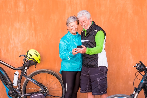 Couple of seniors standing with their two bikes with at red and orang wall at the background - mature two persons using and looking at the same phone