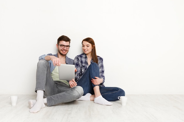 Couple shopping online, making order with digital tablet while sitting at home floor in empty living room