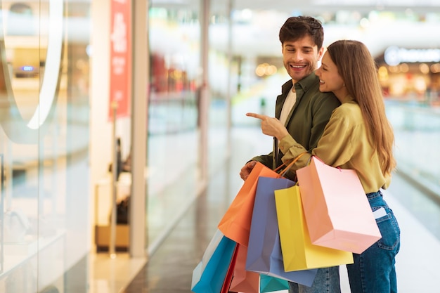 Couple shopping pointing showing clothes in shop window in mall