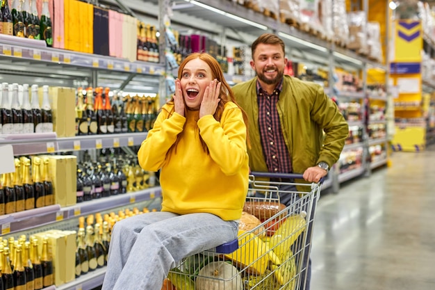 Couple shopping together in grocery supermarket, man carry his redhead girlfriend on cart, they have fun, enjoy time, woman is surprised happy