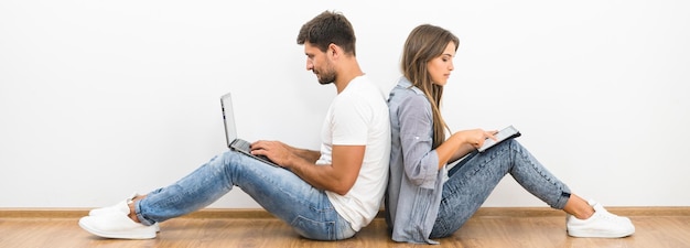 The couple sit with a laptop and a tablet near the empty wall