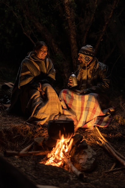 A couple sits around a campfire in the dark, with a blanket over their feet.