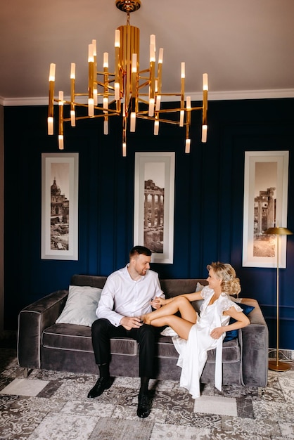A couple sits on a couch in a room with a chandelier hanging above them.