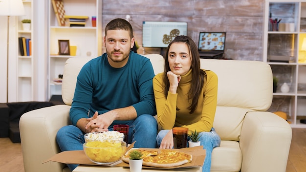 ソファに座ってテレビを見ながらピザを食べながら笑っているカップル。コーヒーテーブルの上のポップコーンとピザ。