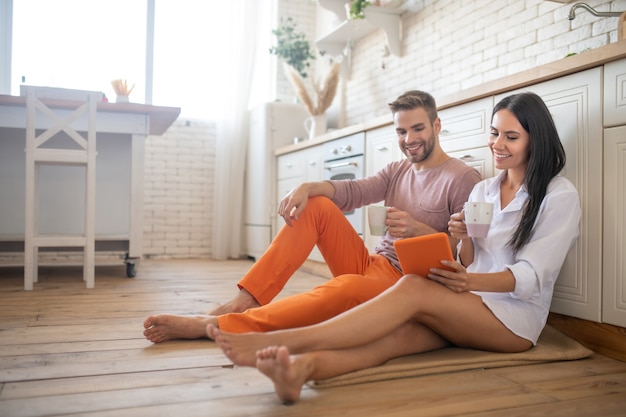 Coppia seduta sul pavimento mentre beve caffè e guarda film su tablet