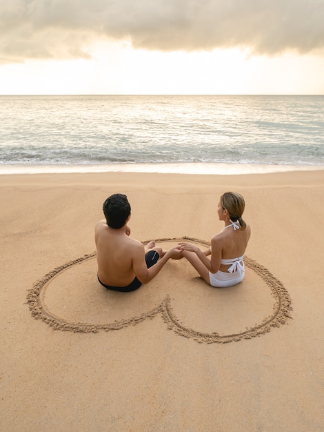 Couple sitting in heart drawing on sand beach relaxing sunbathing.