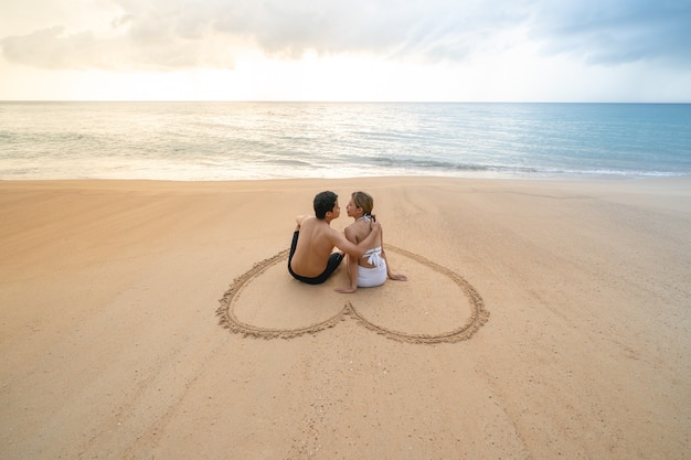 Couple sitting in heart drawing on sand beach relaxing sunbathing.