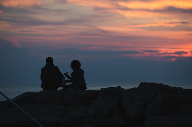 Couple sitting on rocky cliff looking on sunset romantic date summer vacation