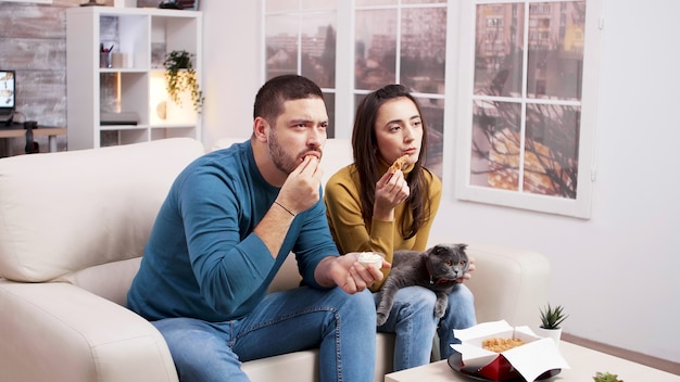 女の子の膝の上で猫と一緒にテレビを見ながらフライドチキンを食べてソファに座っているカップル。ジャンクフードを食べるカップル。