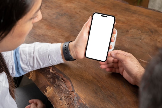 Couple sitting wooden table, looking white blank screen of mobile phone. Vertical smartphone mock up