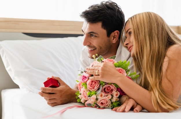 結婚指輪の箱を手で押しながらベッドの上の恋人と幸せな笑顔のカップル