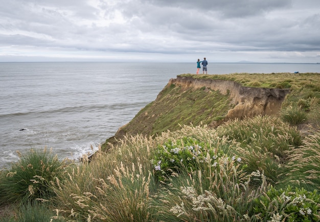 Coppia in piedi sul bordo della scogliera godendo di vista sull'oceano durante il giorno nuvoloso nuova zelanda