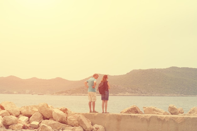 Пара, стоящая на скалистом пляже и смотрящая на море и горы в тонусе
