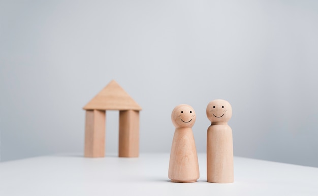 A couple starts a life together with the property real estate investment concept. The happy face wooden human couple standing in front of a wood house miniature on white background, minimal style.