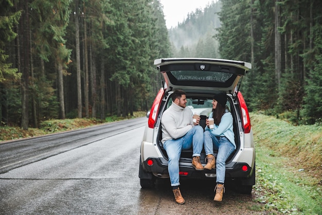 Couple stop to drink hot tea road trip concept sitting in suv\
trunk at road side in mountains forest