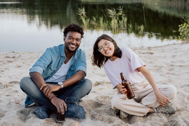 Couple of students friends sitting next to each other on beach\
by lake