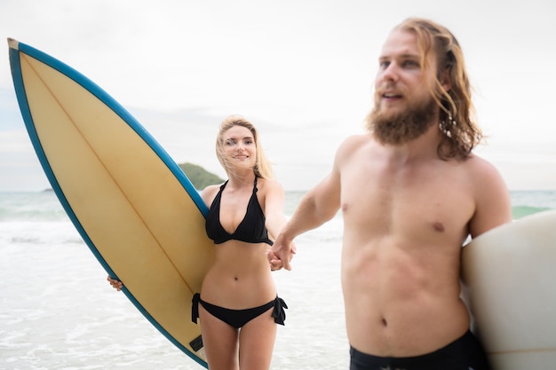サーファーのカップルが手をつないでビーチで互いを眺めている