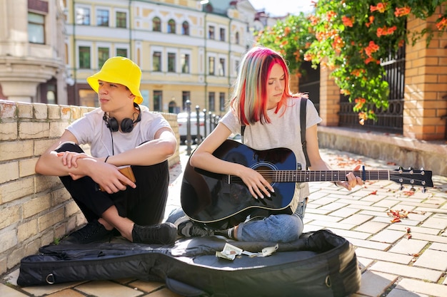 Couple of teenage hipsters street musicians making money by music girl with colored hair singing and playing acoustic guitar Teenagers sitting on sidewalk on city street