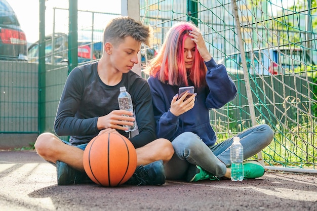 ボール、ライフスタイル、16歳の男の子と女の子が話し、スマートフォンの画面を見てボトルから水を飲むスポーツストリートコートのティーンエイジャーのカップル