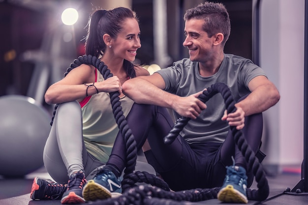 Photo a couple that finished battle ropes workout sit together on a ground inside the gym, looking at each other, smiling.