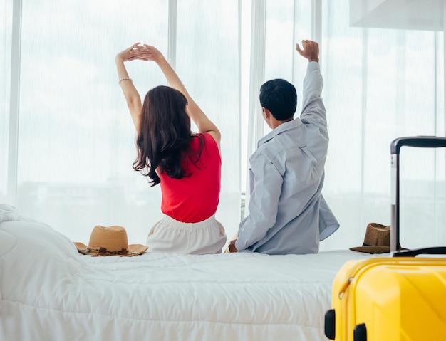 Couple of tourists happy holiday Summer vacation Portrait of back view of young Asian man and woman stretching with relaxing on white bed with suitcase near curtain at the window in hotel room