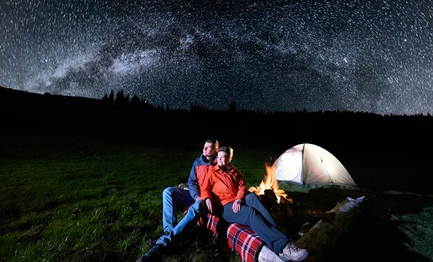 Couple tourists near campfire and tents under night sky full of stars