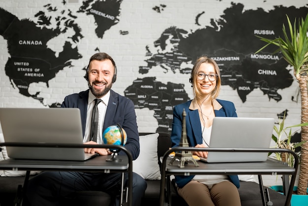 Couple of travel managers working online with laptops and headsets at the agency office with world map on the background