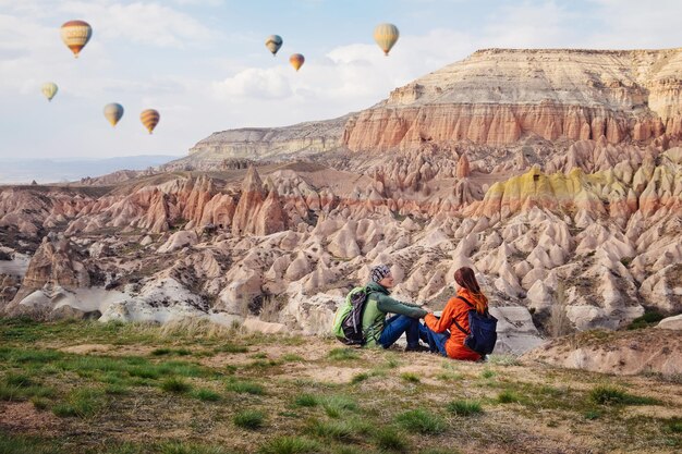 Couple of travelers enjoying landscape of Red valley in Cappadocia near Goreme on the sunset. Man and woman sitting on the cliff of mountains in Turkey.