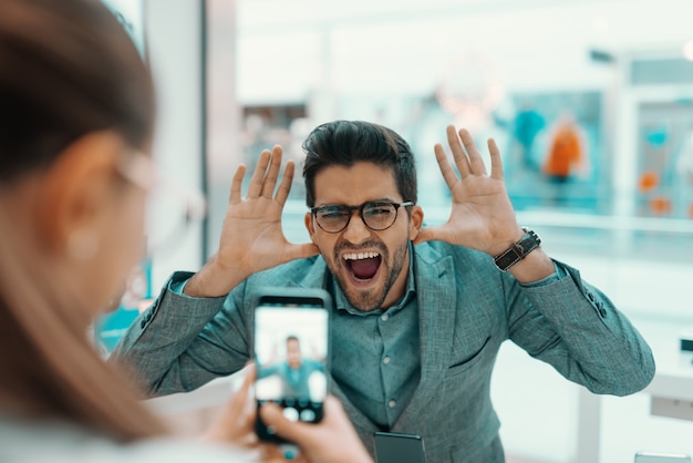 Couple trying out new smart phone in tech store. Woman taking photo of her husband goofing around.