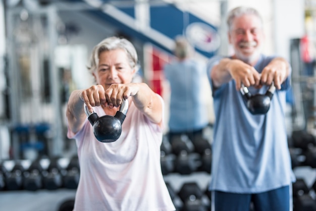 Photo couple of two active and healthy seniors or pensioners or mature people doinng exercise together in the gym holding a dumbbell in their hands and doing squats - fitness lifestyle dieting concept