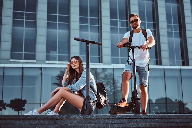 Coppia di due amici attraenti si stanno rilassando vicino a un edificio di vetro con i loro scooter elettrici.