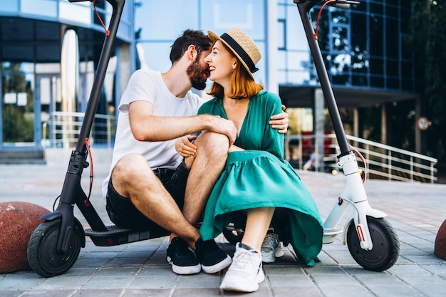 Couple of two attractive people are chilling near the glass building with their electro scooters. Man and woman enjoy vacation