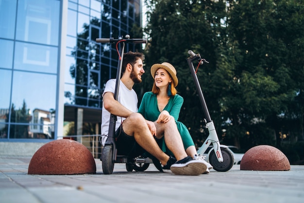 Couple of two attractive people are chilling near the glass building with their electro scooters. Man and woman enjoy vacation