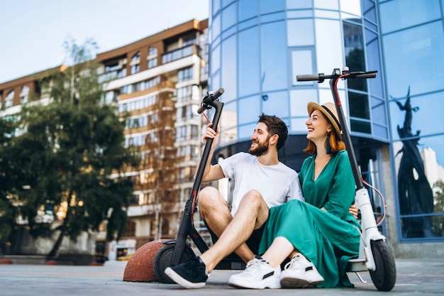 Couple of two attractive people are chilling near the glass building with their electro scooters.